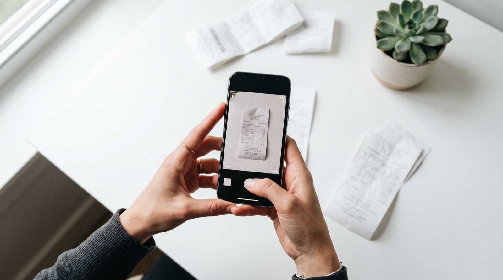 Hands holding a smartphone over a paper receipt mid-scan, with thermal receipts scattered on a white desk and a small plant in the corner