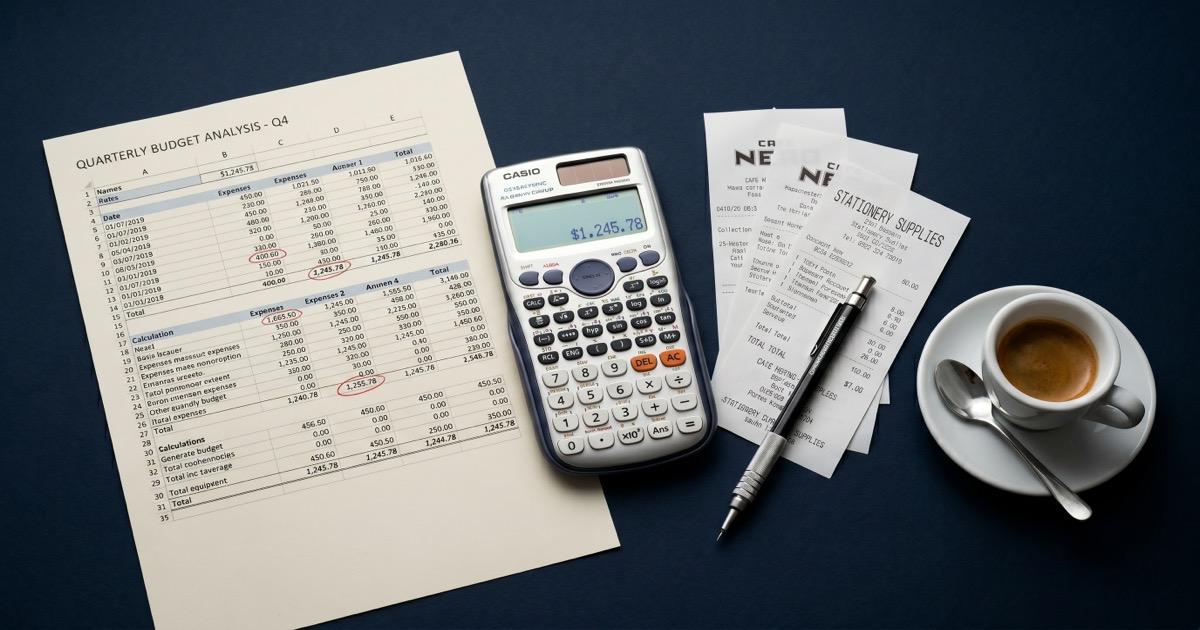 Open laptop showing a revenue dashboard beside an iPhone with an App Store chart, a notebook with calculations, and a coffee on a dark navy seamless backdrop